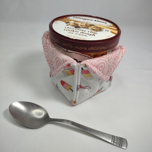 May include: A white and pink fabric ice cream container holder with a zipper closure. The container is decorated with a pattern of pink swirls and ice cream cones. The holder is holding a pint of Häagen-Dazs chocolate chip cookie dough ice cream.