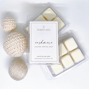 May include: Six white soy wax melts in a clear plastic container. The label reads "PURITY AVE. cashmere cocoa butter. white musk. coconut natural soy wax melts hand poured in MA | 2.75oz/77g"