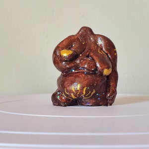 Puede incluir: Una pequeña figurita de cerámica marrón con detalles dorados y purpurina iridiscente. La figurita tiene forma de persona con el vientre redondeado y los brazos cruzados sobre el pecho.