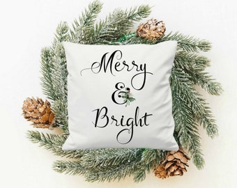 May include: White throw pillow with black text that reads "Merry & Bright" with a small green holly sprig with red berries. The pillow is surrounded by a green pine branch with brown pine cones.
