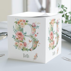 May include: A white square notepad holder featuring a floral design with the letter "B" and the name "Beth". The letter is adorned with pink and peach roses, green leaves, and butterflies. The design is watercolor style.