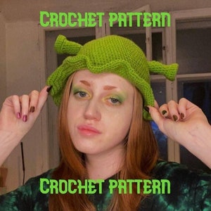 May include: A green crochet hat with two ear-like appendages, resembling a character from a popular animated film. The hat is worn by a person with long red hair and green eyeshadow. The text "CROCHET PATTERN" appears twice in the image.