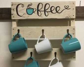Może przedstawiać: Drewniana półka z palety z namalowanym napisem "Coffee". Półka ma pięć haczyków do zawieszania kubków. Na haczykach wisi cztery kubki, dwa turkusowe i dwa białe.