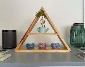 Puede incluir: Una estantería triangular de madera con tres niveles. El nivel superior tiene una pequeña regadera verde con una planta dentro. El nivel intermedio tiene tres pequeños recipientes redondos con patrones coloridos. El nivel inferior está vacío.