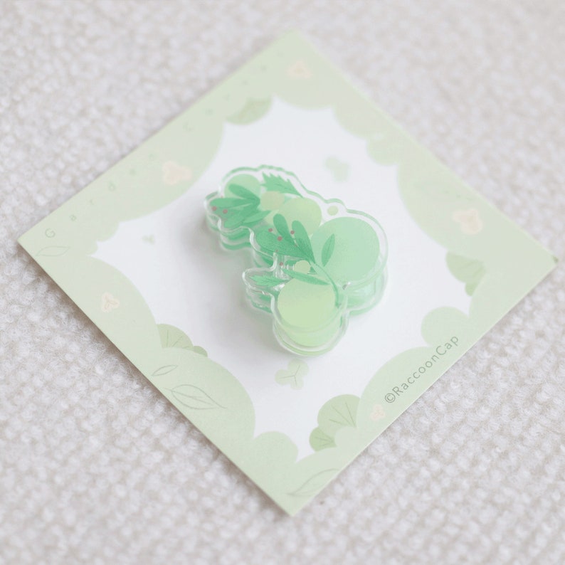 May include: A green and white clear resin pin with a leafy design and the text "@RaccoonCap".