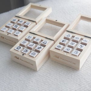 May include: Three light-coloured wooden boxes with hinged glass lids, each containing nine small square rubber stamps. The front of each box is branded with the text RaccoonCap and a small raccoon icon. The stamps feature various black line art icons.