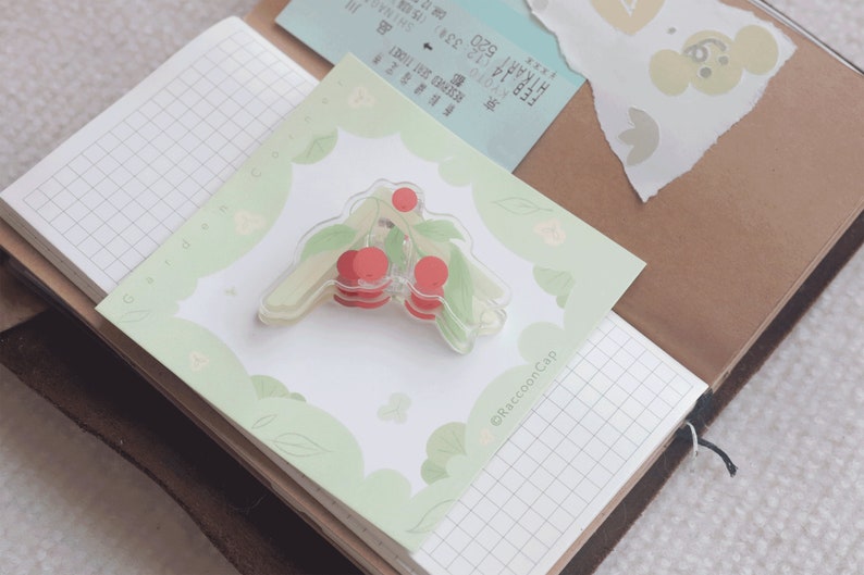 May include: A clear plastic sticker with a cherry design. The sticker is on a green and white background with the text "&copy;Raccoon Cap" in the bottom right corner.