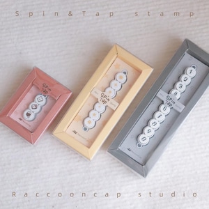 May include: Three Spin & Tap Stamp sets in rectangular boxes. The smallest box is pink with a three-icon stamp. The middle box is yellow with a five-icon stamp. The largest box is gray with a ten-digit number stamp. Each box features the brand name.