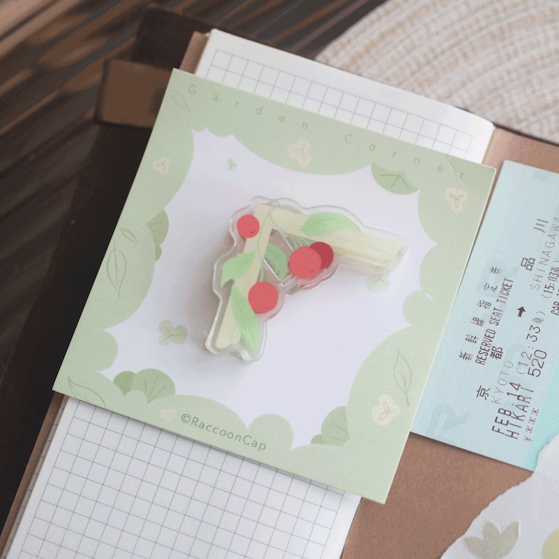 May include: A clear plastic sticker featuring a green and white cherry branch design. The sticker has red cherries and green leaves. The sticker is on a green and white background with the text "Garden Corner" and "@RaccoonCap".