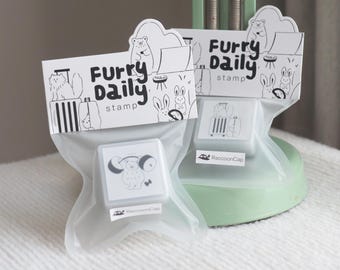 May include: Two clear plastic bags each containing a square stamp. The stamps have a white background with a black illustration of a bear lifting weights and a suitcase. The bags have a white label that reads "Furry Daily stamp" with animal illustrations. The text "RaccoonCap studio" is at the bottom.