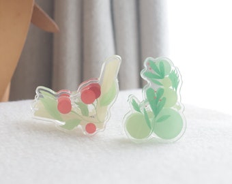 Puede incluir: Dos pinzas para el pelo de acrílico transparente con diseños florales. Una pinza presenta bayas rojas y hojas verdes, mientras que la otra tiene hojas verdes y círculos verde claro.