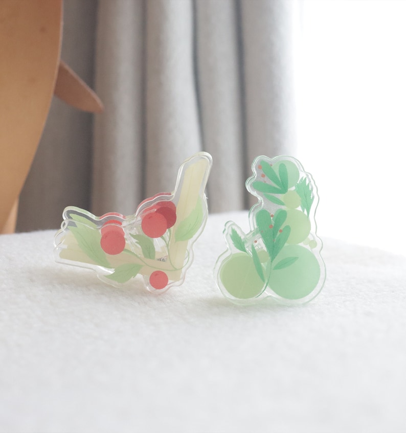 May include: Two clear acrylic hair clips with floral designs. One clip features red berries and green leaves, while the other has green leaves and light green circles.