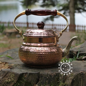 May include: A hammered copper kettle with a wooden handle and a brass spout. The kettle has a lid with a brass knob and is sitting on a tree stump. The text "GALAXY COPPER" is printed on the stump.