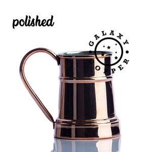 May include: A polished copper mug with a handle. The mug has a rounded bottom and a flared top. The mug is sitting on a white surface. The text "GALAXY COPPER" is printed in black on a white circle above the mug.