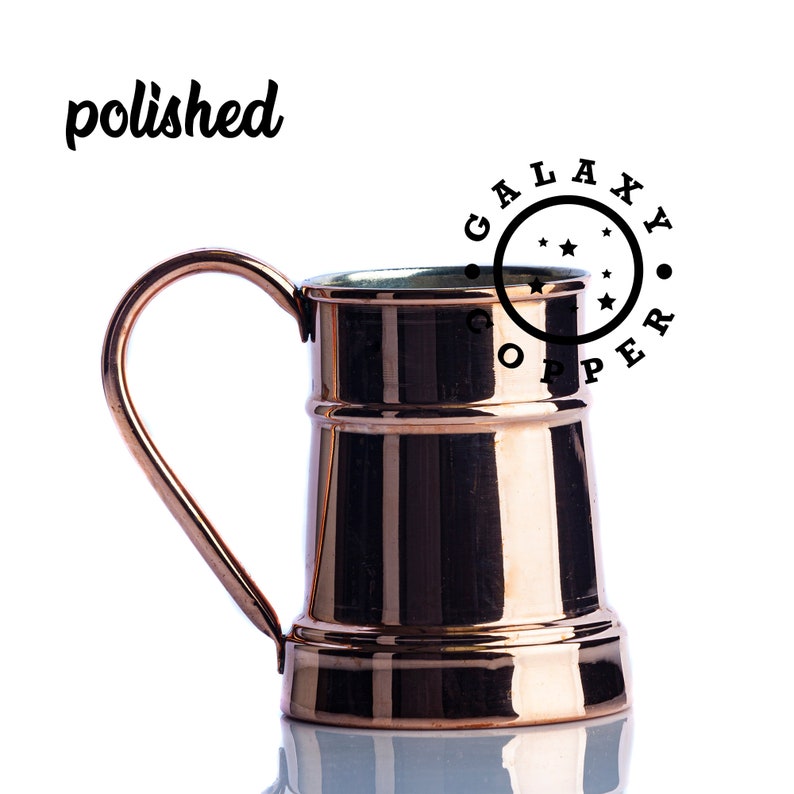 May include: A polished copper mug with a handle. The mug has a rounded bottom and a flared top. The mug is sitting on a white surface. The text "GALAXY COPPER" is printed in black on a white circle above the mug.