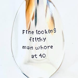 May include: A silver spoon with the words "Fine looking filthy man whore at 40" in black handwritten font. The spoon is oval-shaped and reflects light.