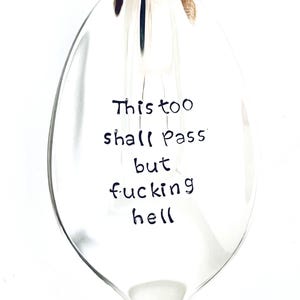 Può includere: Un cucchiaio di vetro trasparente con il testo scritto a mano "This too shall pass but fucking hell" in inchiostro nero. Il cucchiaio ha un lungo manico e una ciotola arrotondata. Lo sfondo è bianco.