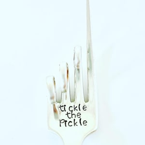May include: A silver fork with the words "tickle the pickle" engraved on the handle. The fork is bent into a hand gesture.