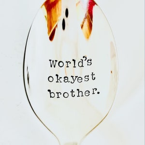 Puede incluir: Una cuchara de plata con las palabras "world's okayest brother." grabadas en ella.