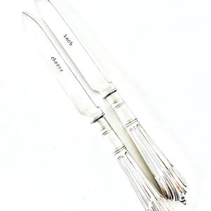 May include: Two silver cheese knives with ornate handles. One knife is labelled "cheese" and the other "knob". The knives are angled against a white background. The handles have a fluted design.