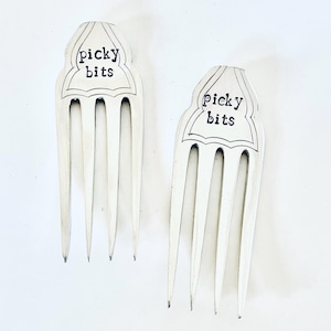 Può includere: Due forcine bianche per capelli con la scritta "picky bits" in corsivo nero. Ogni forcina ha una parte superiore decorativa con un bordo smerlato e quattro rebbi. Le forcine sono su uno sfondo bianco.