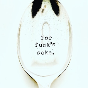 May include: A silver slotted spoon with the phrase "For fuck's sake" stamped in black, centered on the bowl. The spoon has a long handle and is set against a white background.