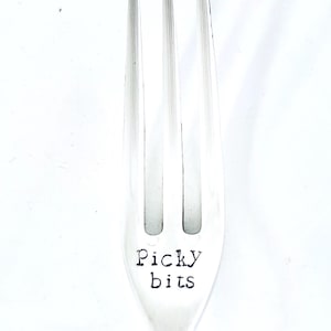 Könnte beinhalten: Eine silberne Gabel mit drei Zinken, gestempelt mit den Worten "Picky bits" in einer Schreibmaschinenschrift. Die Gabel ist vor einem weißen Hintergrund zentriert, wodurch das Design und der Text des Utensils hervorgehoben werden.