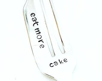 Op de afbeelding: Een zilveren vork met de woorden "eat more cake" in zwarte letters. De vork heeft drie tanden en een gebogen handvat. De achtergrond is wit.