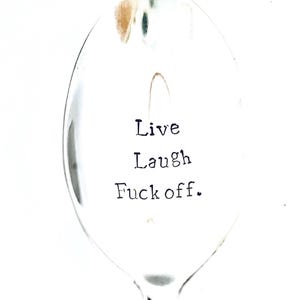 Puede incluir: Una cuchara de plata con el texto "Live Laugh Fuck off." grabado en la parte delantera.