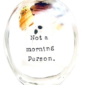 May include: A clear glass wine glass with the text "Not a morning person." in black, centered within the glass. The glass has a rounded bowl and a stem.
