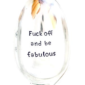 以下が含まれることがあります： 「Fuck off and be fabulous」の文字が黒色で書かれた透明なスプーン。スプーンのボウルの中央に配置されています。スプーンは金と銀の色合いの反射面を持っています。