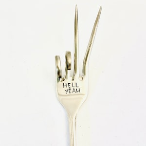 Puede incluir: Un tenedor de plata doblado en forma de mano haciendo el dedo medio. El tenedor tiene las palabras "Hell Yeah" grabadas en el mango.