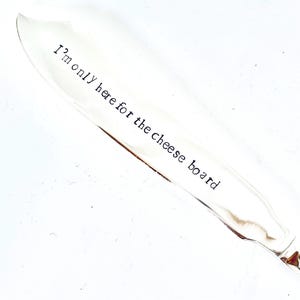 May include: A silver cheese knife with a rounded tip. The text "I'm only here for the cheese board" is printed on the blade in black. The knife is set against a white background.