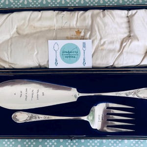 Puede incluir: Un juego de servir plateado, que incluye un cuchillo y un tenedor, presentado en una caja negra con interior azul. El cuchillo tiene el texto "All you need is love... and a slice of life." El tenedor dice "Yeah!"