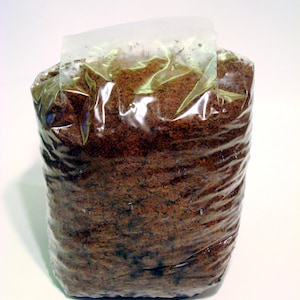 May include: A close-up of a brown, loose, granular material packaged in a clear plastic bag. The bag is sealed at the top and the material is visible through the plastic.