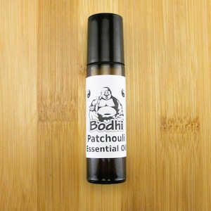 May include: A brown glass bottle with a black roll-on top. The bottle has a white label with the text "Bodhi Patchouli Essential Oil" and a black and white image of a laughing Buddha.
