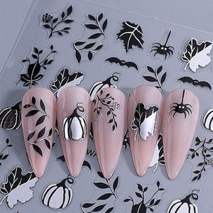 May include: Nail art stickers featuring black and silver designs. The stickers include leaves, pumpkins, and spiders. The nail art is applied to pink, almond-shaped nails. The background is a clear plastic sheet.