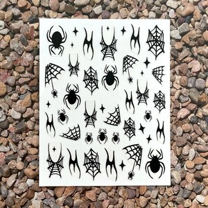 May include: A sheet of black and white nail art stickers featuring spiders, spiderwebs, and star shapes. The stickers are arranged on a white background, perfect for Halloween or spooky-themed nail designs.
