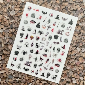 May include: A sheet of Halloween-themed nail art stickers. The stickers feature various spooky designs, including skulls, bats, spiders, and tombstones. The text "WG672" is printed on the sheet. The stickers are in black, white, and red.