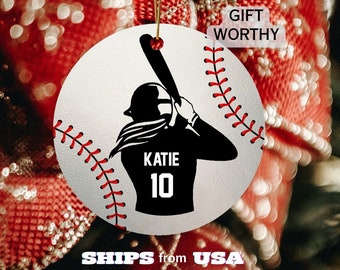 May include: White round ornament with a black silhouette of a baseball player swinging a bat. The player is wearing a baseball cap and jersey with the name "KATIE" and number "10" on it. The ornament has a baseball stitch design around the edge.