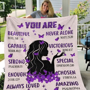 May include: A soft, pink and white blanket with a silhouette of a woman with purple butterflies. The blanket has inspirational text with bible verses, including "You Are Beautiful", "Never Alone", "Capable", "Victorious", "Strong", "Special", "Enough", "Chosen", "Always Loved", and "Amazing".