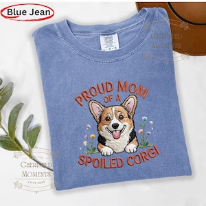 Puede incluir: Una camiseta azul de cuello redondo con un bordado de un perro Corgi y el texto PROUD MOM OF A SPOILED CORGI en naranja. La prenda tiene una etiqueta Comfort Colors en el cuello interior y una textura de tela con aspecto desgastado.