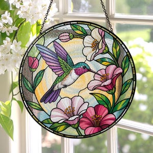 May include: A round stained glass window panel featuring a hummingbird in flight, surrounded by pink and white flowers and green leaves. The bird has purple, green, and blue feathers. The panel is framed in black and hangs from a chain.