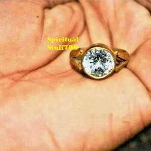 Könnte beinhalten: Ein goldener Ring mit einem großen, klaren, runden Edelstein. Der Ring befindet sich an der Hand einer Person.