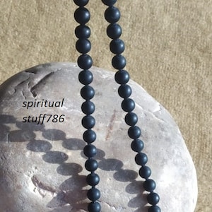 May include: A black beaded necklace made of matte black beads. The necklace is lying on a light brown rock.