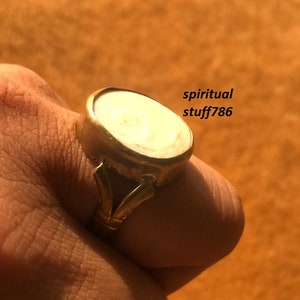 Może przedstawiać: Złoty pierścionek z białym owalnym kamieniem osadzonym w oprawie typu bezel. Pierścionek ma ozdobny pasek z wzorem w postaci zwijającego się elementu.