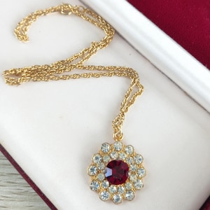 May include: A gold-tone chain necklace featuring a circular pendant. The pendant has a large, faceted deep red centre stone surrounded by a single halo of small, clear, round-cut white rhinestones set in a gold-tone metal frame.