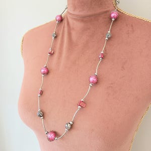Può includere: Una collana lunga caratterizzata da un'alternanza di perle sferiche rosa striate, piccoli distanziatori in metallo argentato e accenti rettangolari rosa irregolari. Le perle sono infilate su una sottile catena argentata in un motivo.