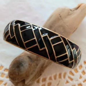 May include: A rigid bangle bracelet featuring a glossy black enamel surface inlaid with a geometric pattern of thin gold-toned metal lines. The design consists of irregular rectangular and angular shapes that wrap around the entire outer surface.