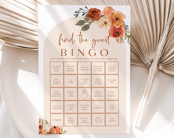May include: A printable bridal shower game card with an orange, white, and brown floral design. The card has the title "Find the Guest Bingo" and includes a grid of squares with various prompts for guests to find someone who matches the description.